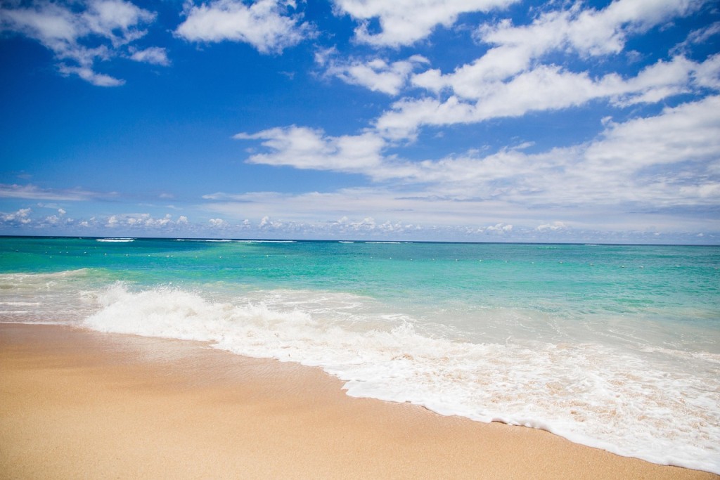 Sandstrand mit blauem Meer bei strahlendem Himmel mit vereinzelten, weißen Wolken.